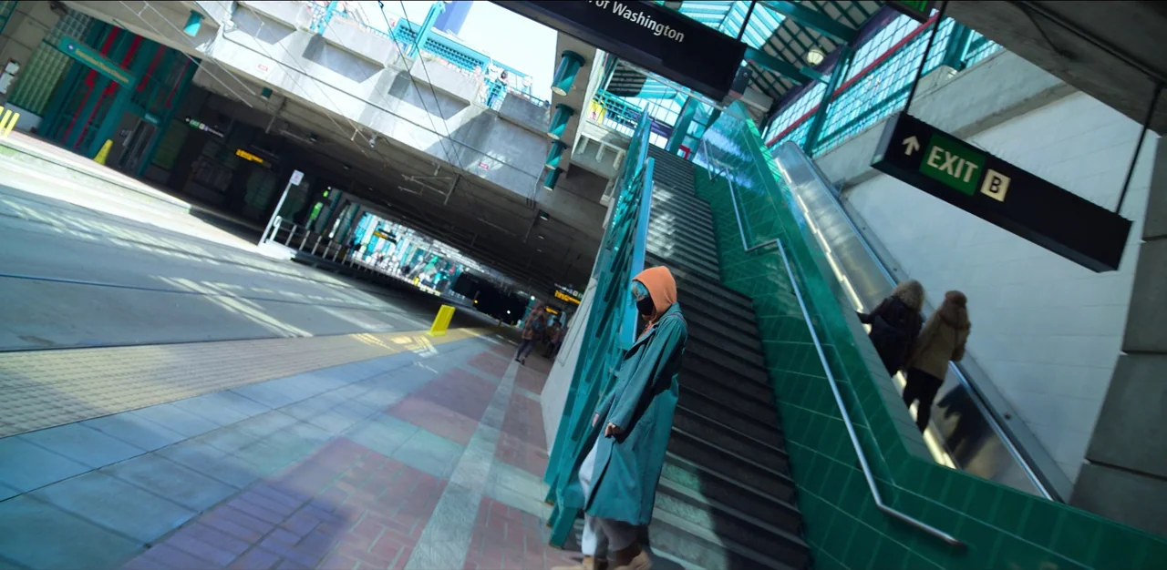Kimi (2022) movie still: fullbody — An outdoor transit station features concrete structures and green ceramic tile accents. A person in a teal coat…