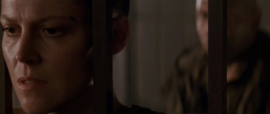 Alien 3 (1992) movie still: closeup — A dimly lit industrial space defined by vertical dark iron bars. A woman stares past the metal bars as a…