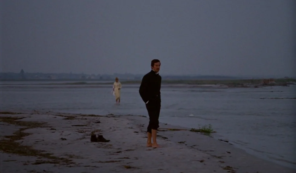 Elvira Madigan (1967) movie still: wide — A desolate, sandy shoreline at dusk under a uniform, overcast gray sky. A man in a dark suit stands…