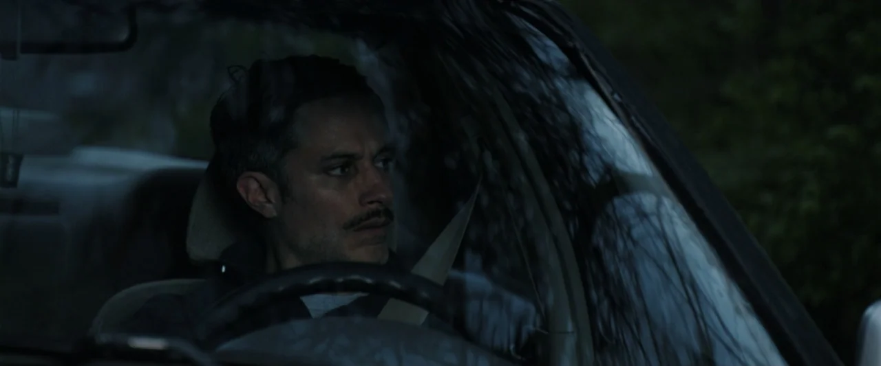 Holland (2025) movie still: medium — A dark vehicle interior with the man visible through the windshield and driver's side window. The man is…
