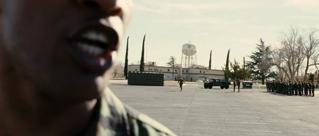 Jarhead movie still: wide — An open military base parade ground with a water tower in the background. A soldier in the foreground yells while a…