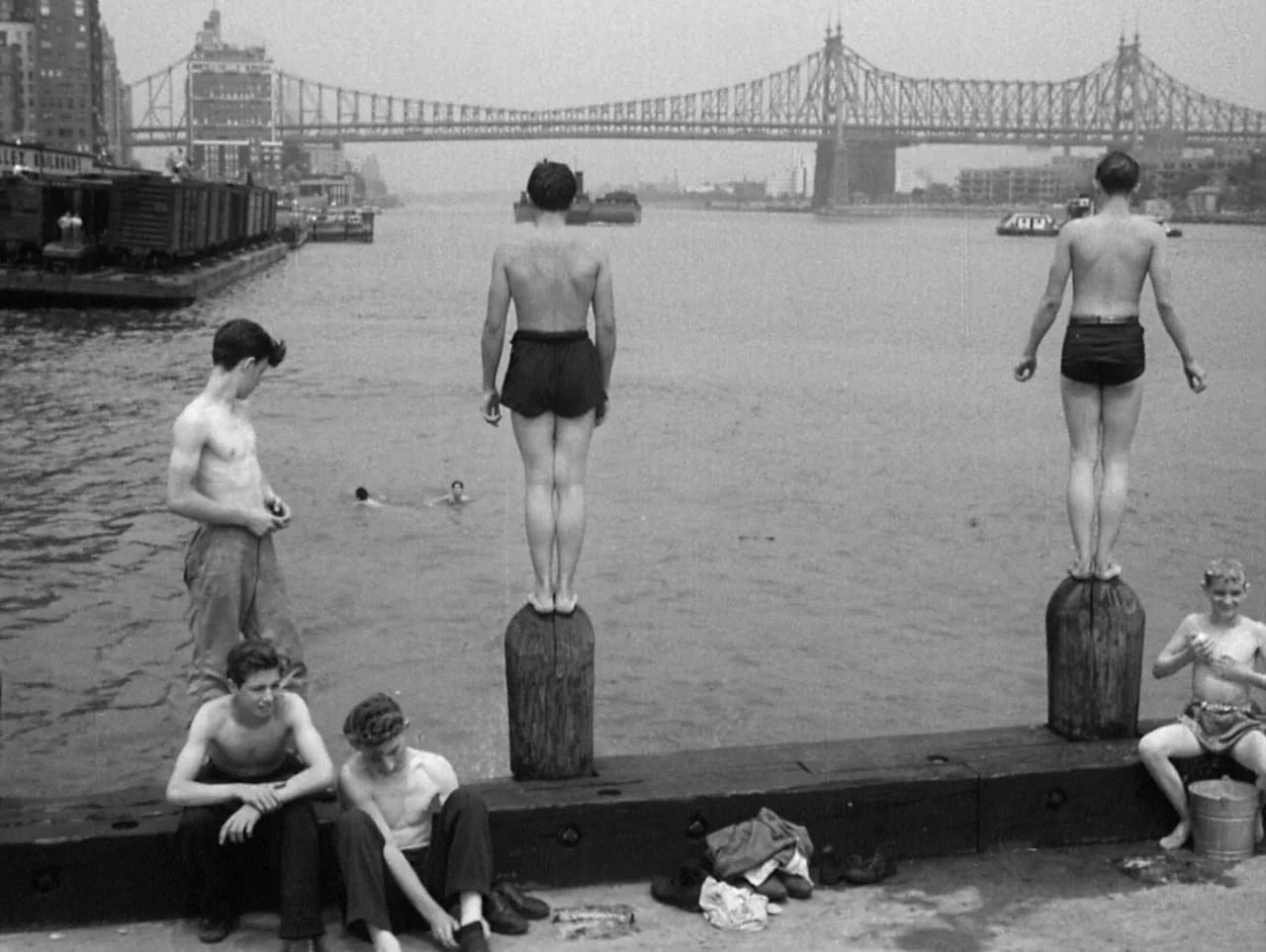 The Naked City (1948) movie still: wide — A group of teenage boys congregate on a wooden pier overlooking a wide river. The Queensboro Bridge spans…