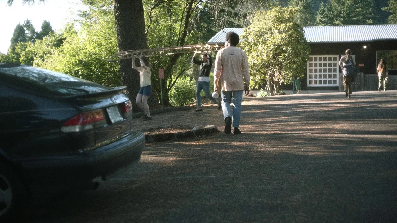 Showing Up (2022) movie still: wide — A paved lot sits in front of a wood-sided building with a metal roof. Four individuals walk across the lot…