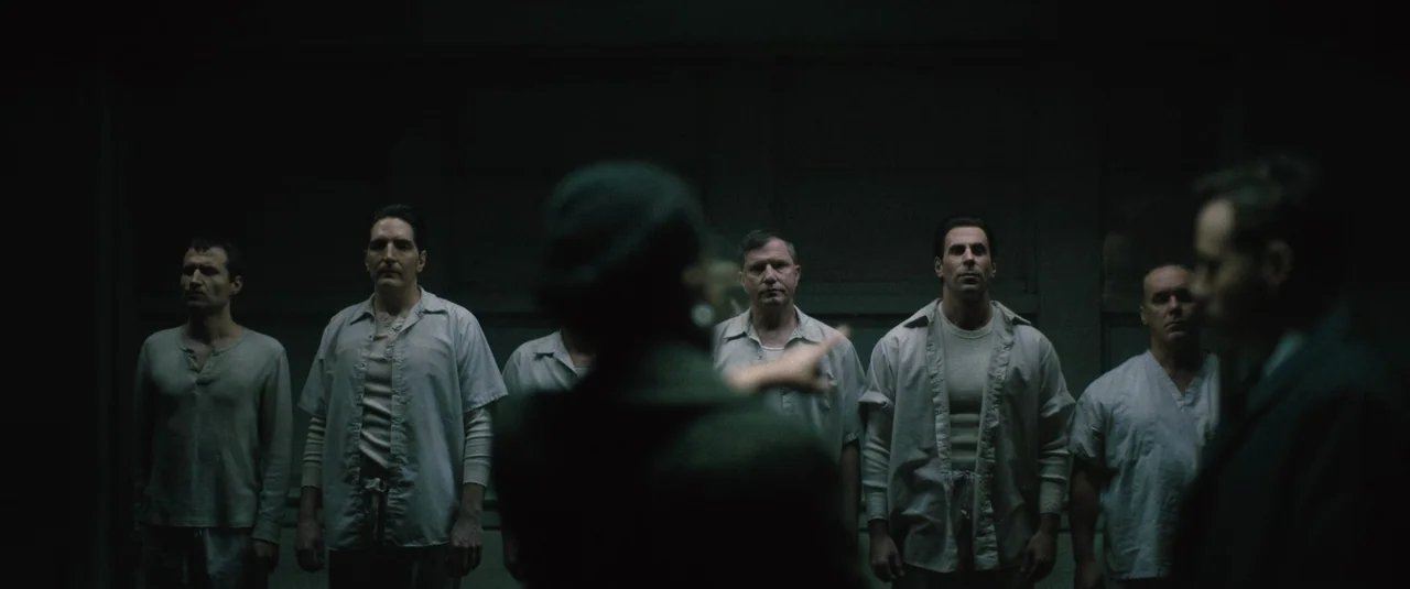 Boston Strangler (2023) movie still: medium — A dark, dimly lit institutional ward features rows of men in uniform hospital shirts. One man in the…