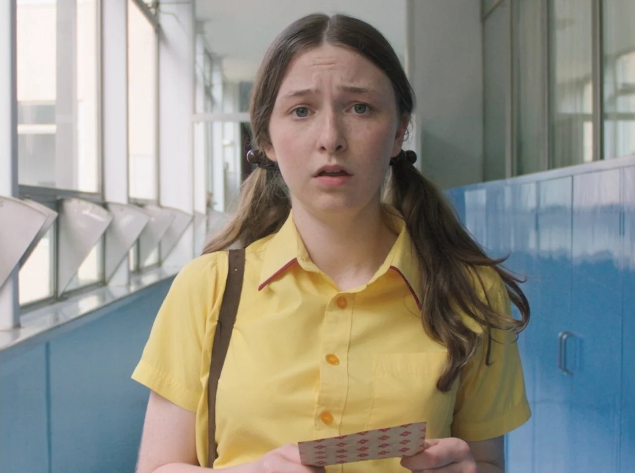 Girl Asleep (2015) movie still: medium — A school hallway with blue lockers and large glass windows on the left side. A teenage girl in a yellow…