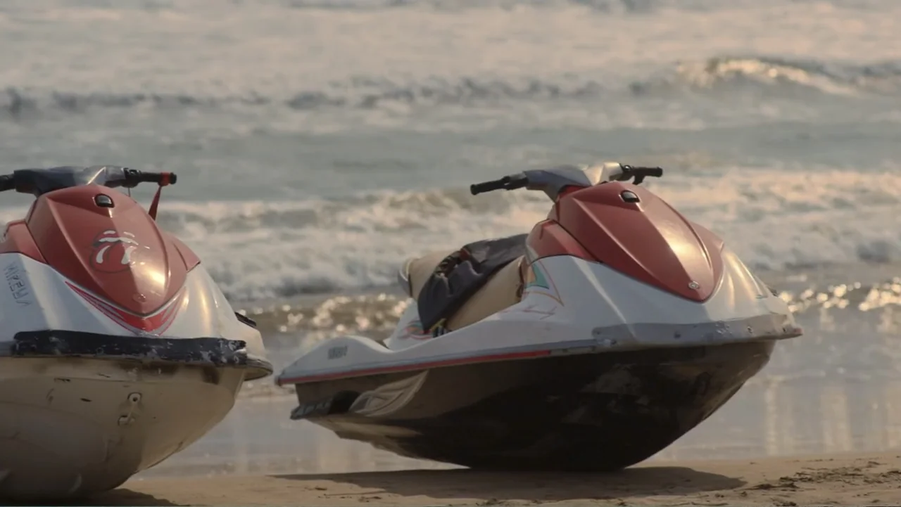 Double Pregnant 2 (2022) movie still: medium — Two white and red jet skis sit parked on a sandy beach shore. The tide laps at the sand behind the…