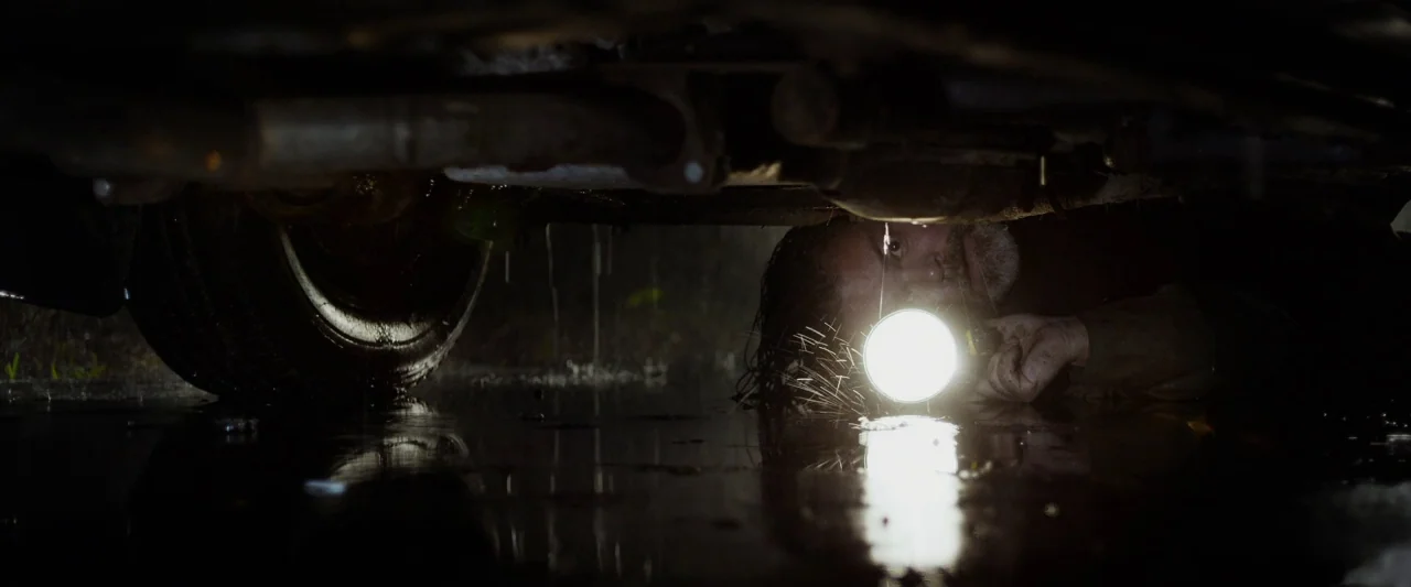 The Monster (2016) movie still: medium — A man lies on his side on wet asphalt under the chassis of a vehicle. He holds a bright flashlight toward…