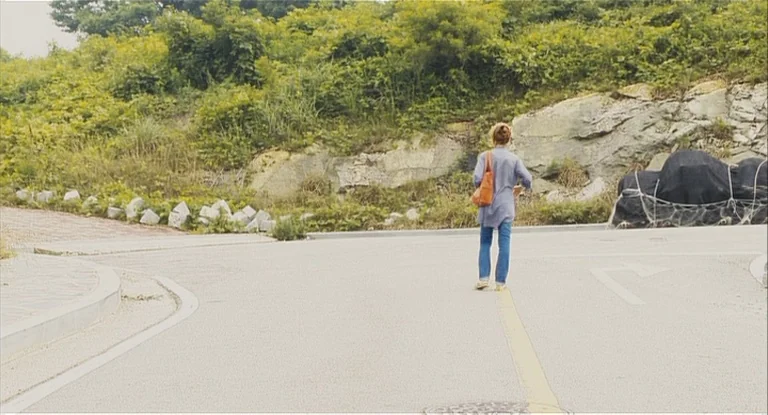 In Another Country (2012) movie still: wide — A paved asphalt road stretches toward a green, rocky hillside covered in wild vegetation. A woman…