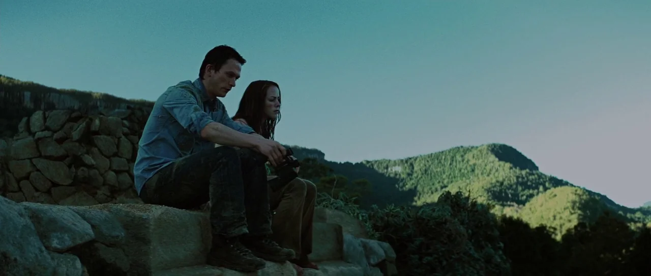 The Ruins (2008) movie still: medium — A man and woman sit on stone steps overlooking a lush green mountain range. They are resting together on the…