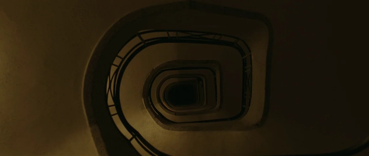 Bullhead (2011) movie still: establishing — A spiraling staircase descends into a dark, void-like center within a dimly lit building. The structure…