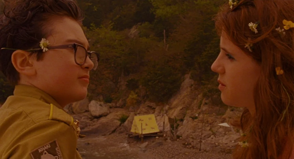 Moonrise Kingdom (2012) movie still: medium — A rocky beach cove features a small yellow tent positioned near the water's edge. A teenage boy and…