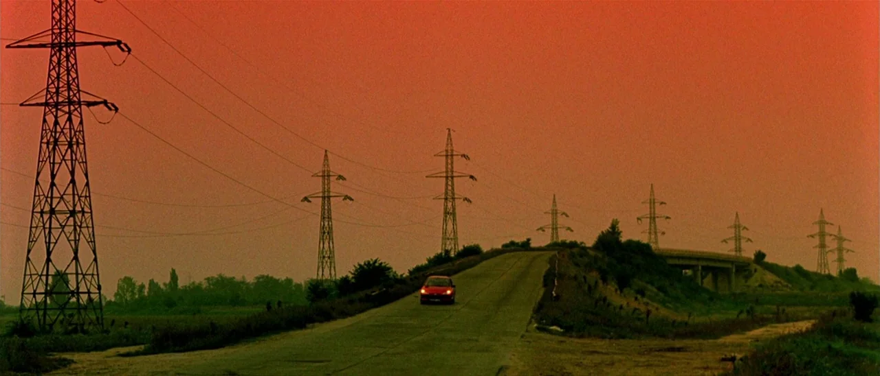 Haute Tension (2003) movie still: wide — A rural asphalt road cuts through a flat landscape under an orange-tinted evening sky. Multiple tall steel…