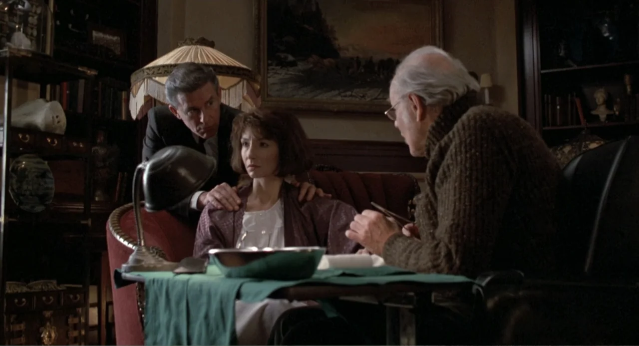 Dead of Winter (2025) movie still: medium — A dimly lit Victorian study features dark wood furniture, bookshelves, and a framed painting. Three…