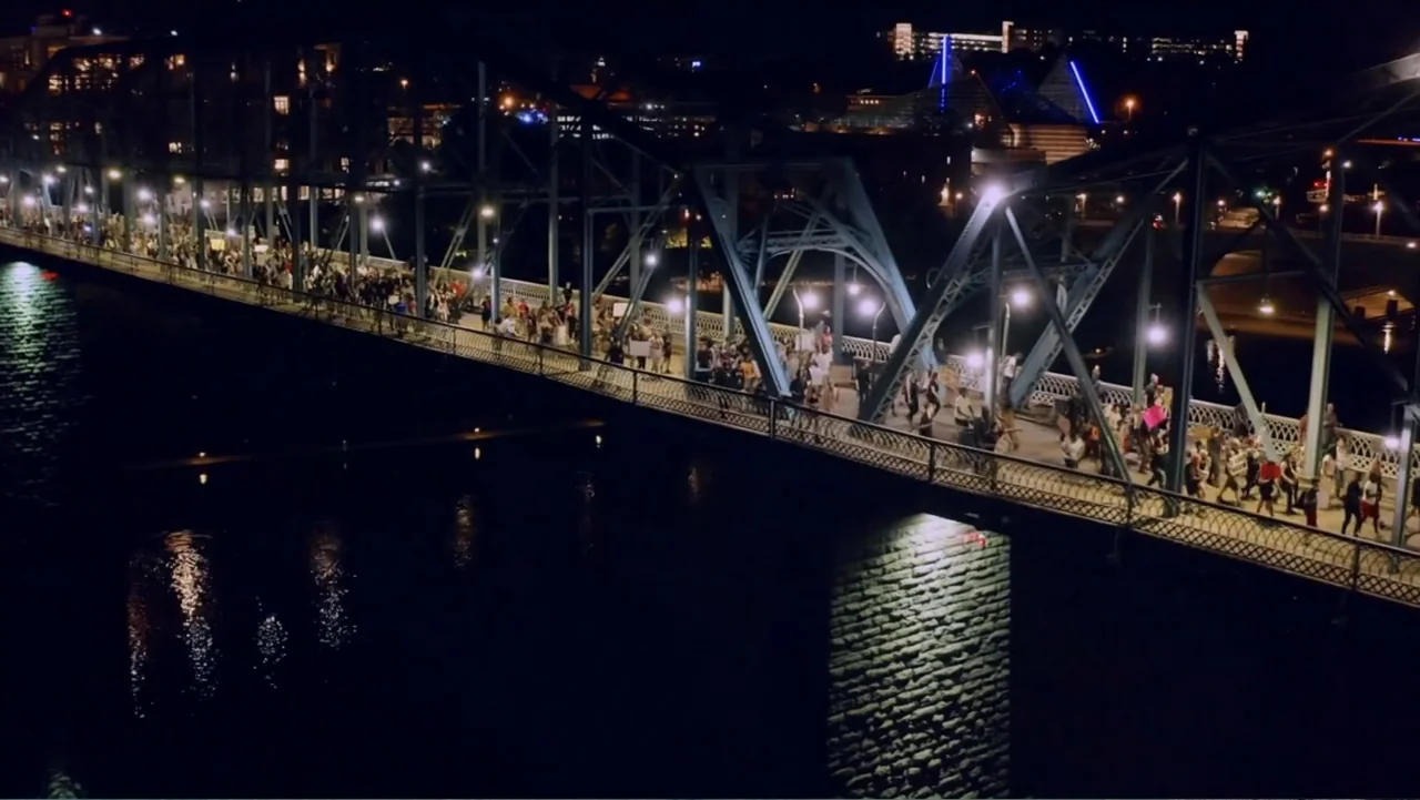 Between the World and Me (2020) movie still: wide — A metal truss bridge spans a dark body of water at night. A large crowd of people walks across…