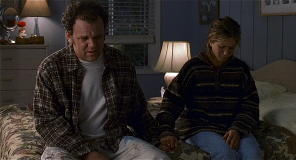 The Good Girl (2002) movie still: medium — A dimly lit bedroom features floral bedding and blue wall paneling. A middle-aged man and a woman sit…