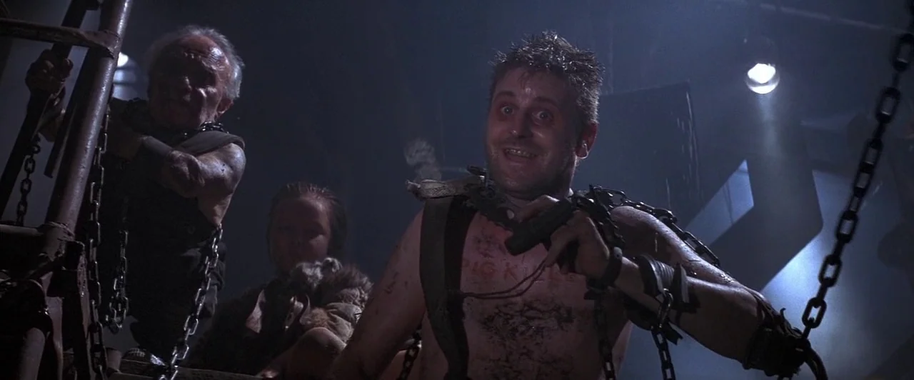 Mad Max Beyond Thunderdome (1985) movie still: medium — A dimly lit room filled with thick heavy metal chains and industrial hardware. Three…