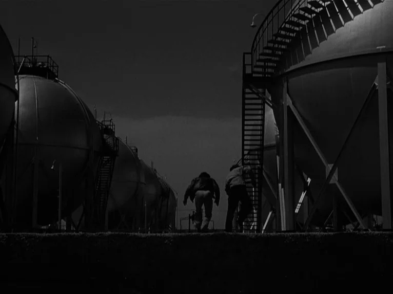 White Heat (1949) movie still: wide — A dark industrial site contains several massive spherical metal tanks and staircase structures. Two men run…