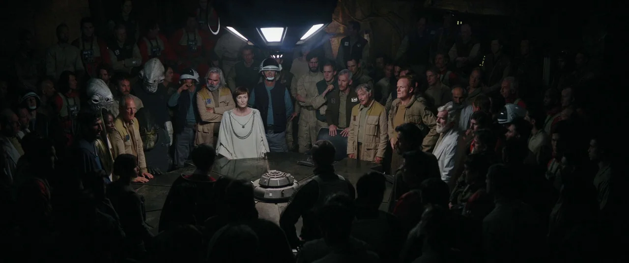 Rogue One: A Star Wars Story (2016) movie still: wide — A dimly lit industrial room features a large central table surrounded by many people. A…