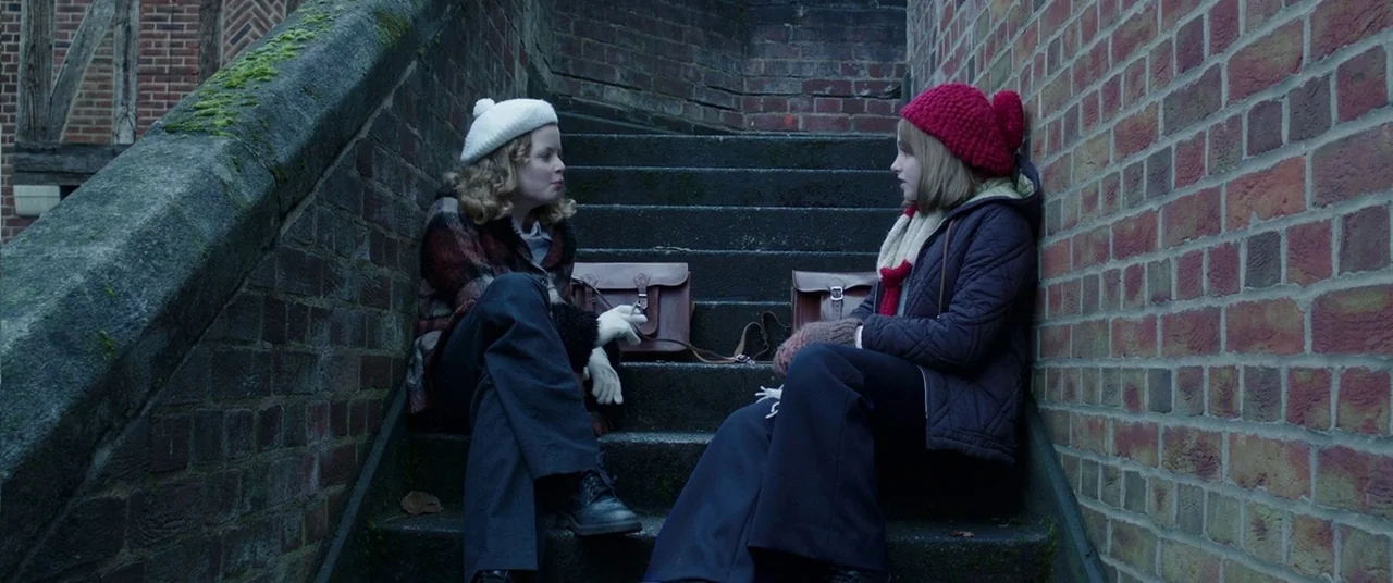 The Conjuring 2 (2016) movie still: fullbody — Two teenage girls sit on stone steps bounded by red brick walls. They engage in conversation while…