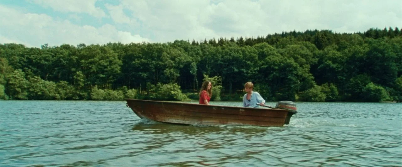Adoration (2019) movie still: wide — A wooden motorboat travels across a calm lake lined with dense evergreen trees. The two occupants sit in the…