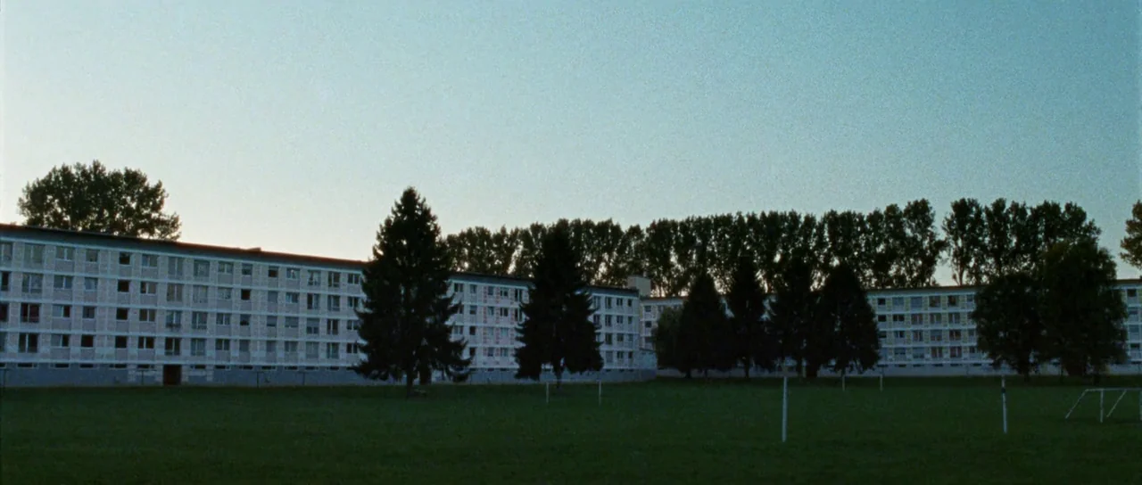 Alléluia (2014) movie still: establishing — A long, multi-story residential apartment block stretches across the frame behind a grass field. Dark…
