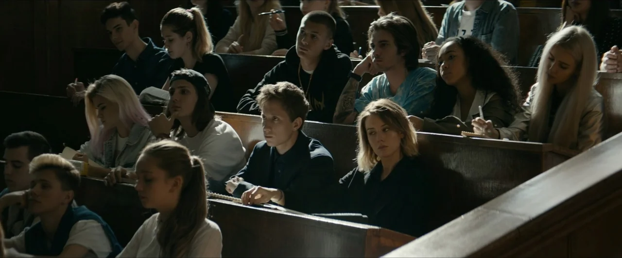 Invasion (2020) movie still: medium — A tiered wooden lecture hall with students sitting in rows at desks. The students observe the front of the…