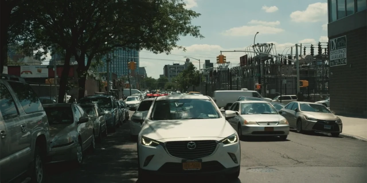 Equal Standard (2021) movie still: wide — An urban thoroughfare features parked and driving vehicles under a clear blue sky. Electrical…