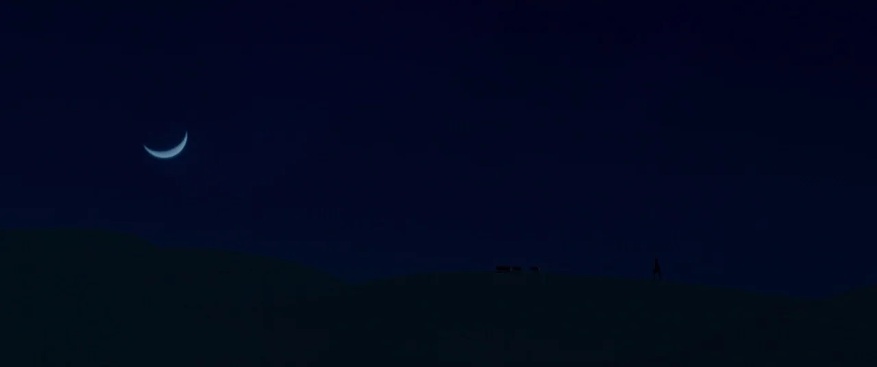 Siberia (2019) movie still: wide — A dark night landscape features rolling sand dunes under a pale crescent moon. A solitary figure walks across the…