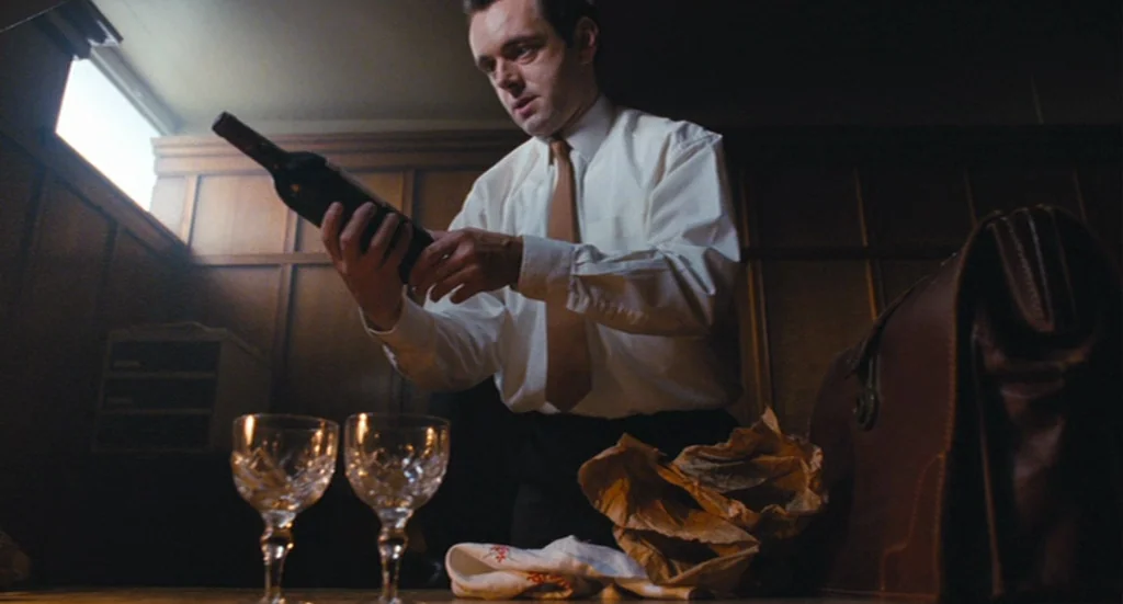 The Damned United (2009) movie still: medium — A man stands in a wood-paneled room near a small window with heavy shadows. He inspects a wine bottle…