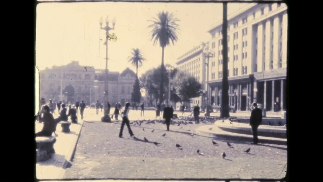 Transfers (2024) movie still: wide — A spacious urban plaza features tall palm trees and large neoclassical architecture in daylight. Numerous…