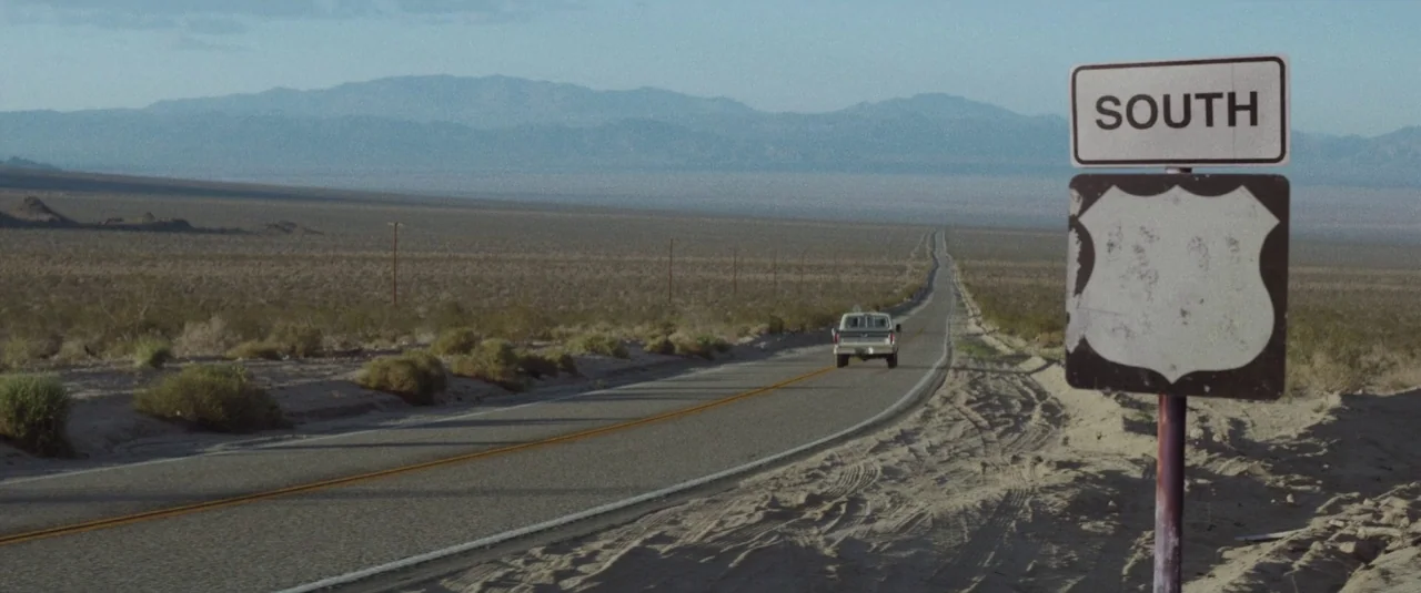 Southbound (2015) movie still: wide — An arid desert highway stretches into the hazy mountain range under a clear sky. A lone white pickup truck…