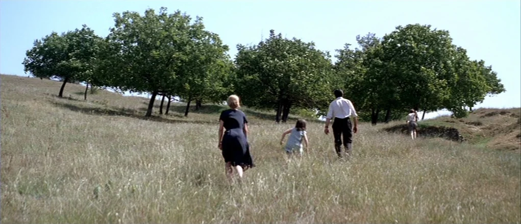 The Banishment (2007) movie still: fullbody — A group walks up a gentle hill covered in dry tall grass toward trees. The terrain is an open grassy…