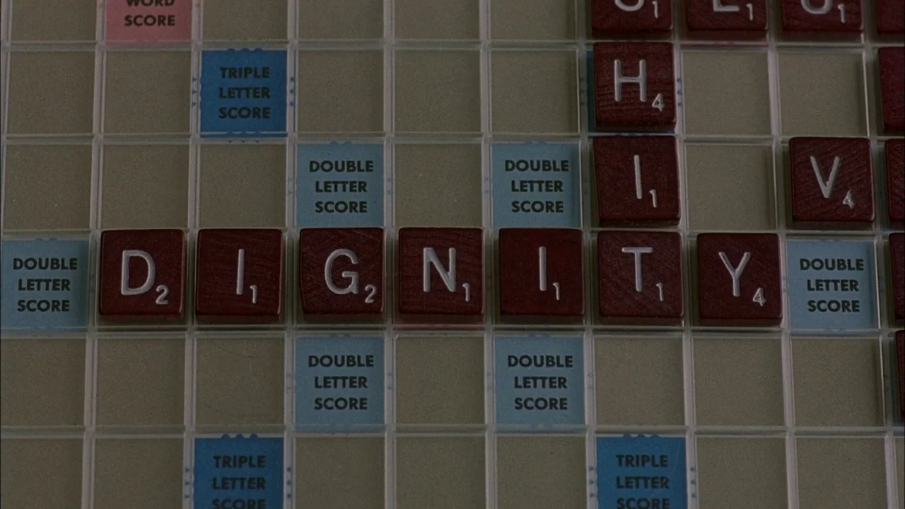 Whore (1991) movie still: closeup — A close-up view of a Scrabble board with maroon plastic letter tiles placed flat. The letters spell the word…