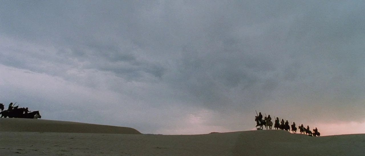 Renegade (2004) movie still: wide — A wide expanse of rolling sand dunes under a heavy, overcast dusk sky. A long line of mounted riders in…