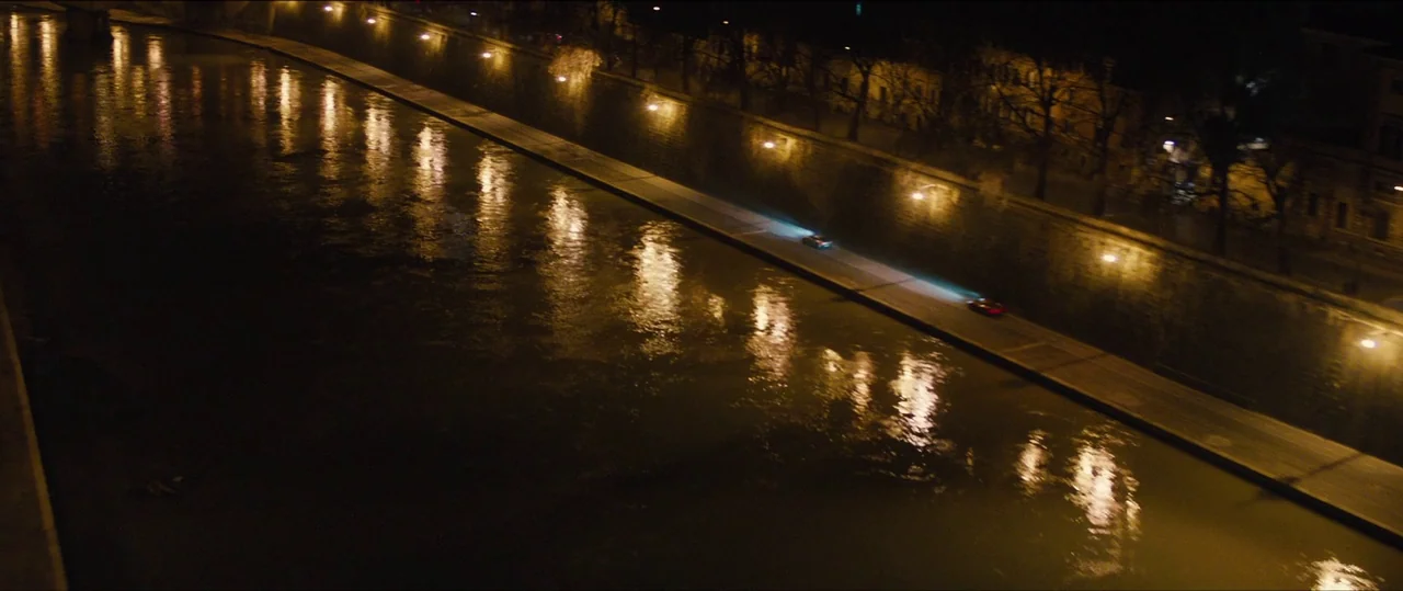 Spectre (2015) movie still: wide — An urban river at night with reflections of golden streetlights on the water. Two vehicles travel in sequence…