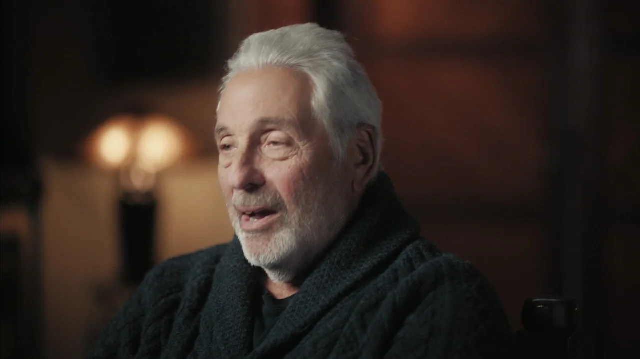 Faye (2024) movie still: medium — An elderly man is seated in a dimly lit, out-of-focus indoor space. He wears a dark sweater and is speaking while…