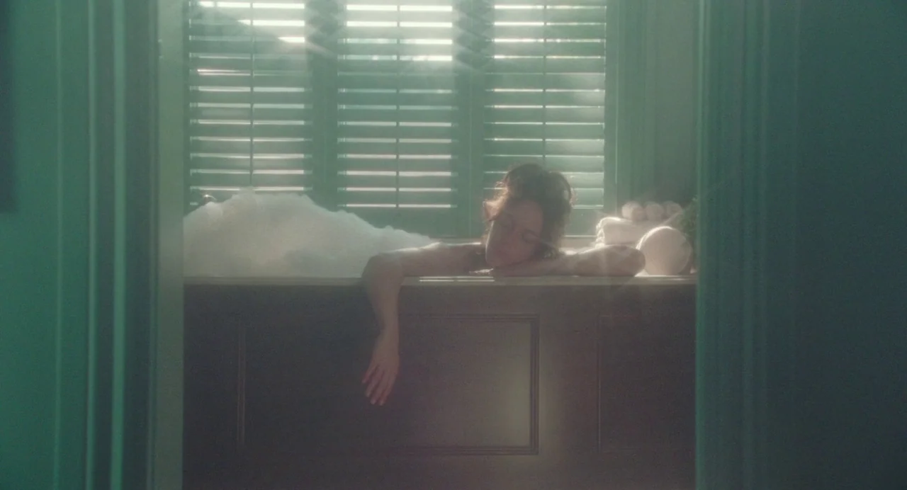 Babysitter (2022) movie still: medium — A bathroom features a tub set against a window with closed green shutters. A woman with closed eyes rests in…