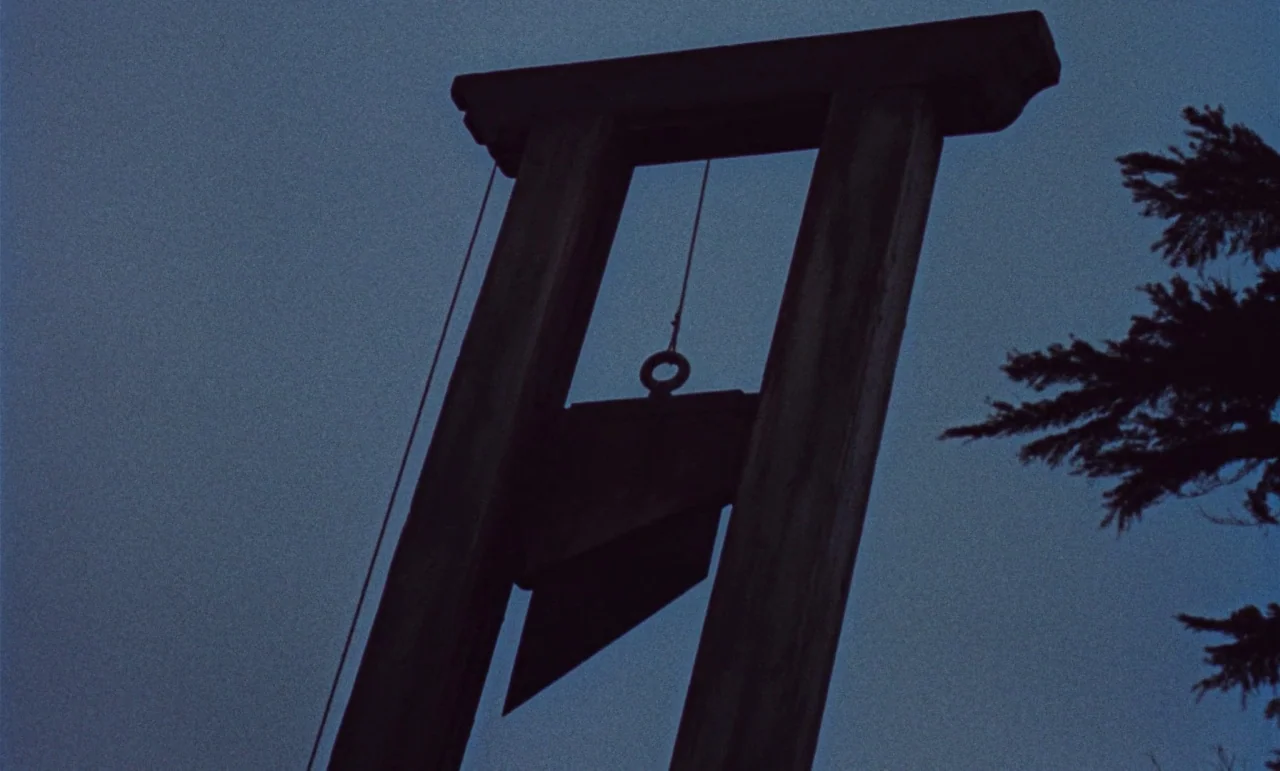 The Revenge of Frankenstein movie still: medium — A wooden guillotine frame stands prominently against a dark, featureless evening sky. The blade is…