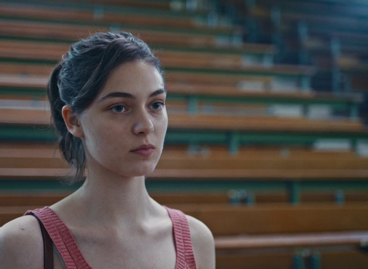 Happening (2021) movie still: medium — An indoor space features rows of wooden stadium benches blurred in the background. A young woman stands in…