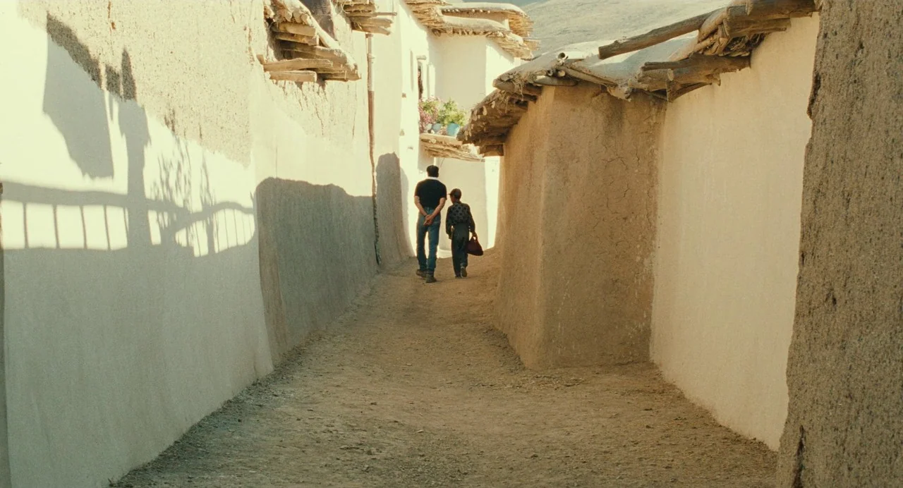 The Wind Will Carry Us (1999) movie still: wide — A narrow alleyway enclosed by high textured earthen walls with protruding wooden ceiling beams. An…