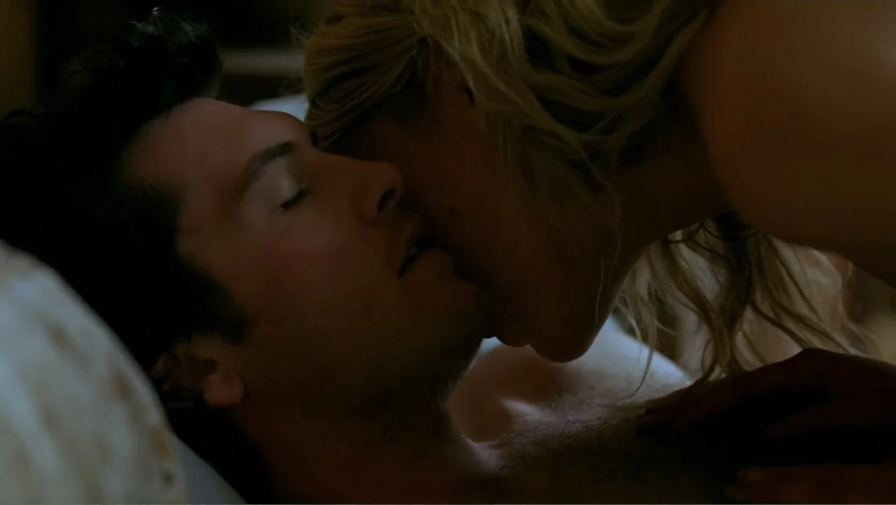 Emily (1976) movie still: closeup — A dark bedroom interior with the subjects lying in bed on white pillows. A woman leans over a man with her hair…