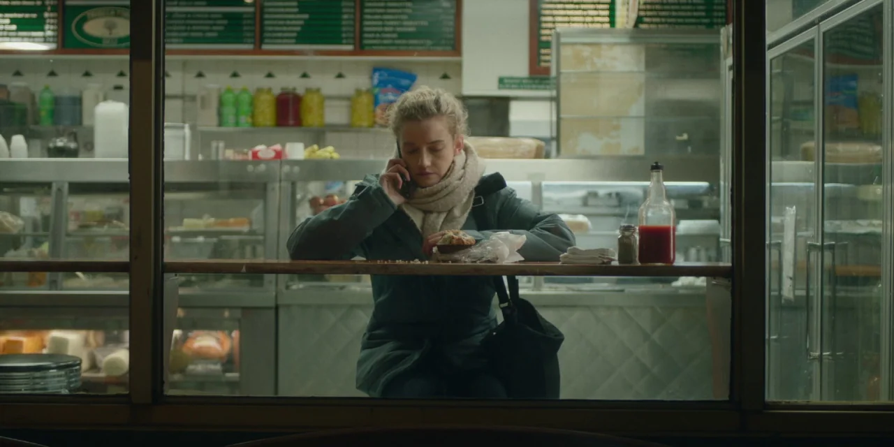 The Assistant (2019) movie still: medium — A woman sits behind a glass partition inside a well-stocked local deli at night. She holds a smartphone…