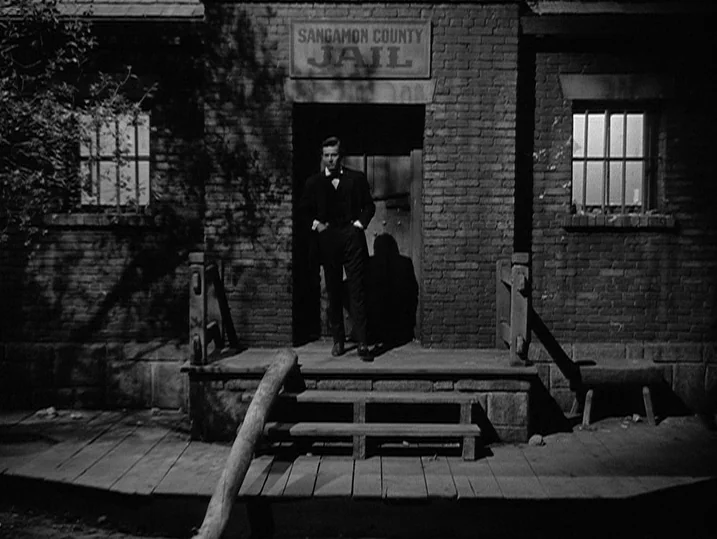 Young Mr. Lincoln (1939) movie still: fullbody — A brick jail building with barred windows and a wooden porch at night. A man stands at the doorway…