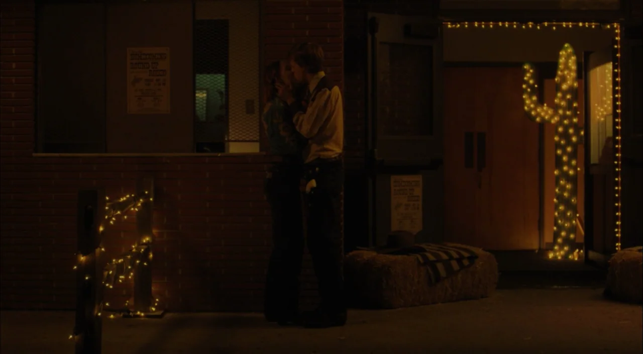 Lady Bird (2017) movie still: fullbody — The brick facade of a school features posters for a rodeo event. A couple stands near the building doors…
