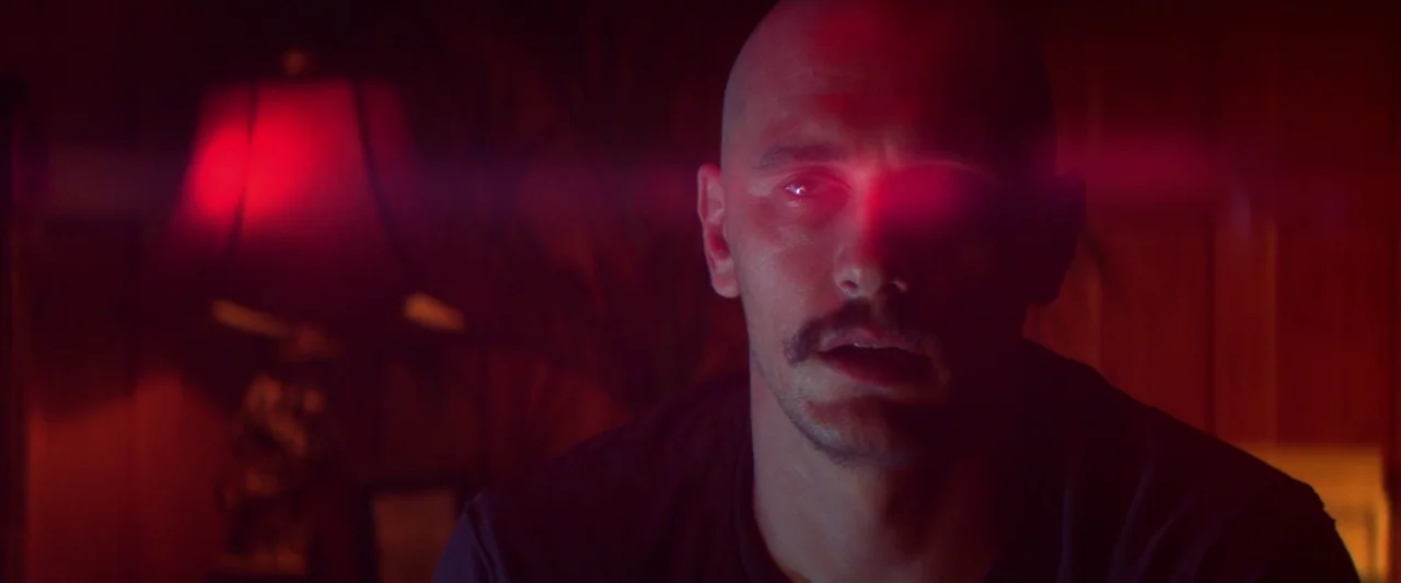 Zeroville (2019) movie still: medium — A dark, dimly lit domestic space contains a single bright red lampshade in the background. A man stands in…