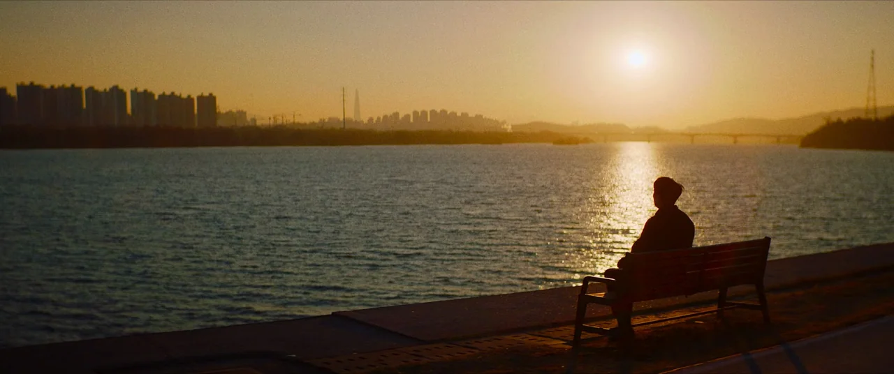 The Divine Fury (2019) movie still: wide — A riverside pathway at golden hour with a distant city skyline under a clear sky. A person sits alone on…