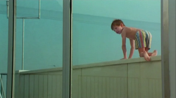 Code Unknown (2000) movie still: medium — A modern balcony with a pale stone ledge and glass sliding doors reflects the sky. A young boy in colorful…