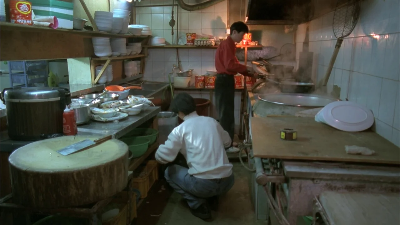 The Day a Pig Fell Into the Well (1996) movie still: wide — A cluttered commercial food preparation area features stacked ceramic bowls and large…