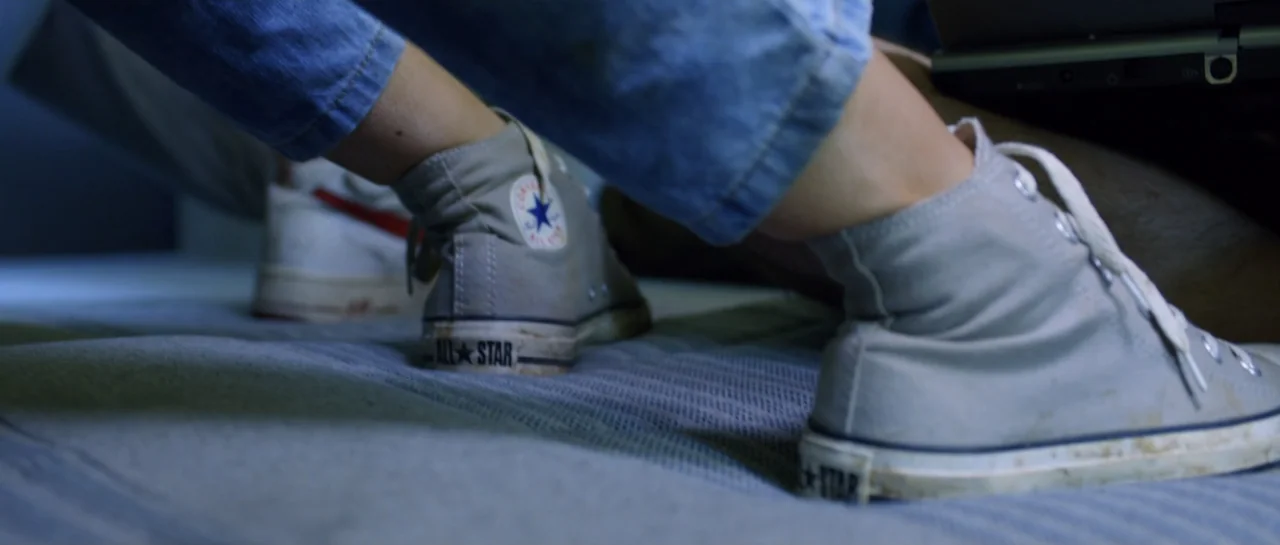 La presencia (2012) movie still: closeup — A dimly lit room focuses on two sets of feet resting on a mattress. The subjects wear high-top sneakers…