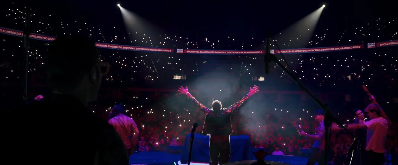 Making of: F*CKS NEWS (2025) movie still: fullbody — A dark, expansive indoor concert arena features a central stage illuminated by powerful beams.…